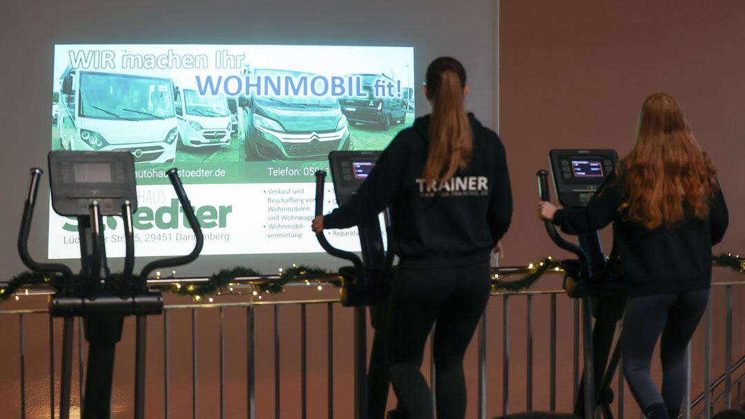 Trainierende im Fitnessstudio Fox Training in Lüchow, im Hintergrund läuft ein lokaler Display-Spot (Autohaus Stoedter) auf einer großen Bildschirmfläche.