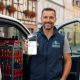 Dienstleister steht vor Servicefahrzeug und zeigt Smartphone mit Online-Profil.